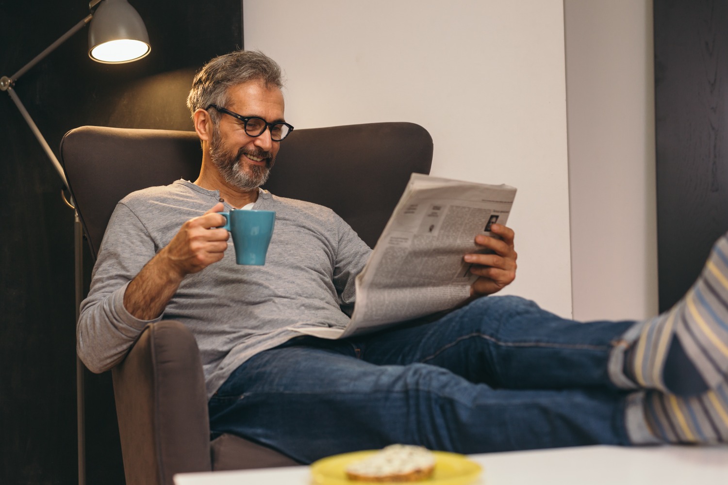 A man wearing glasses sits on a chair, enjoying a cup of coffee while reading a newspaper.