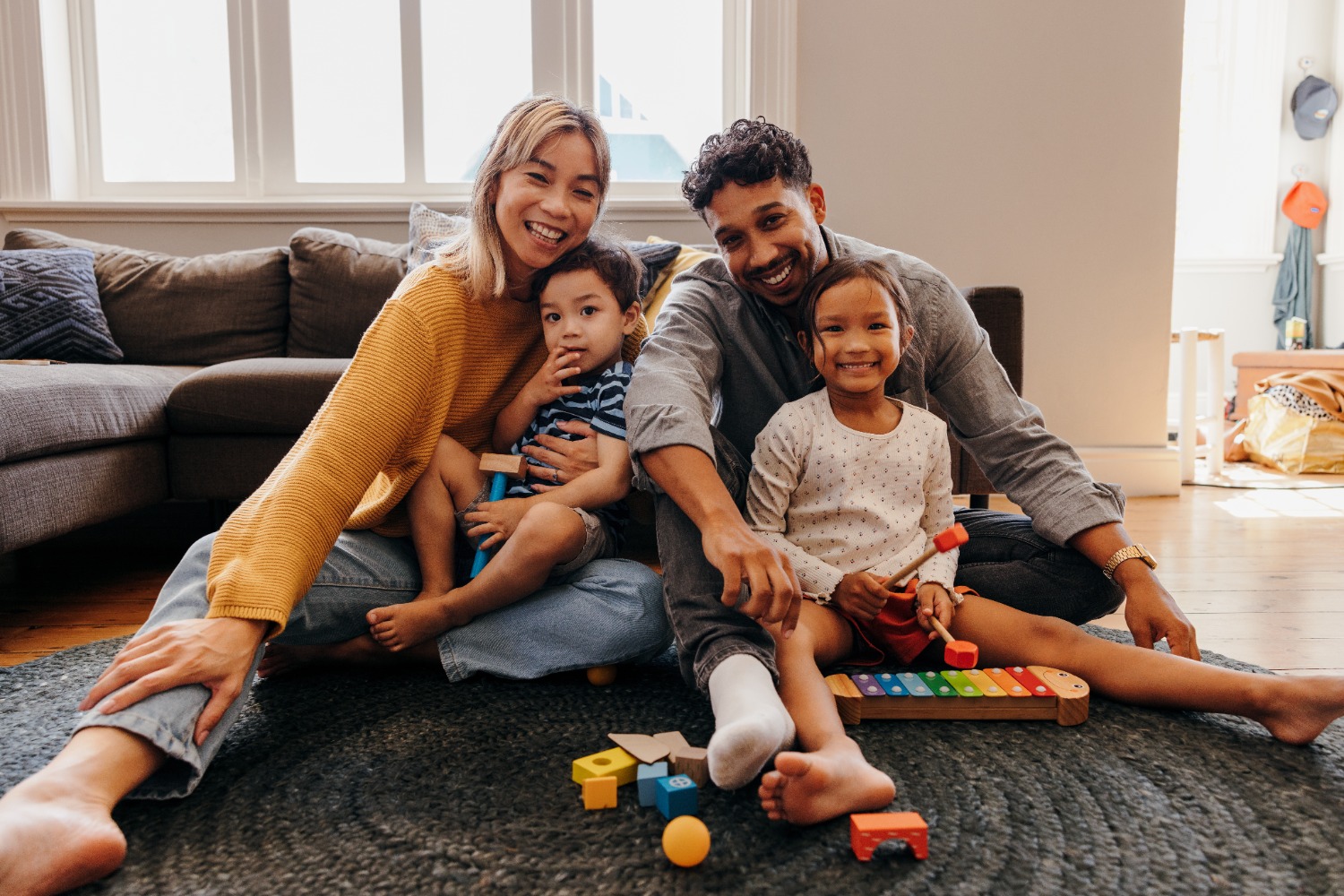 Two adults and two children sitting on the floor in a cozy living room, playing with colourful building blocks and a wooden toy xylophone.