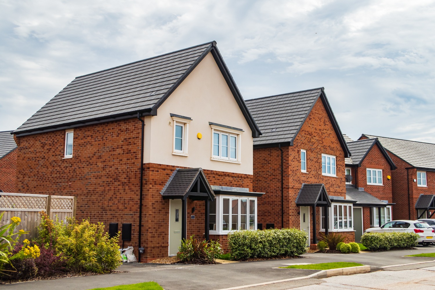 Modern residential area with detached houses featuring brick facades and dark tiled roofs, each with a driveway and well-maintained lawn under a partly cloudy sky.