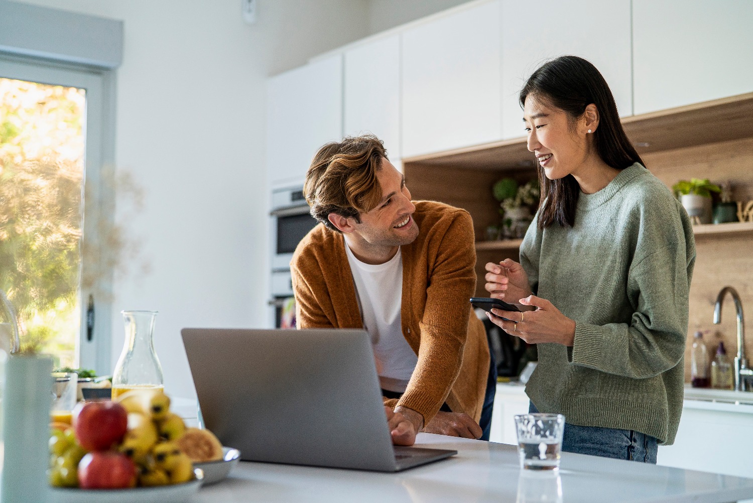 Adult couple looking for online mortgage deals on laptop and smart phone at kitchen counter.